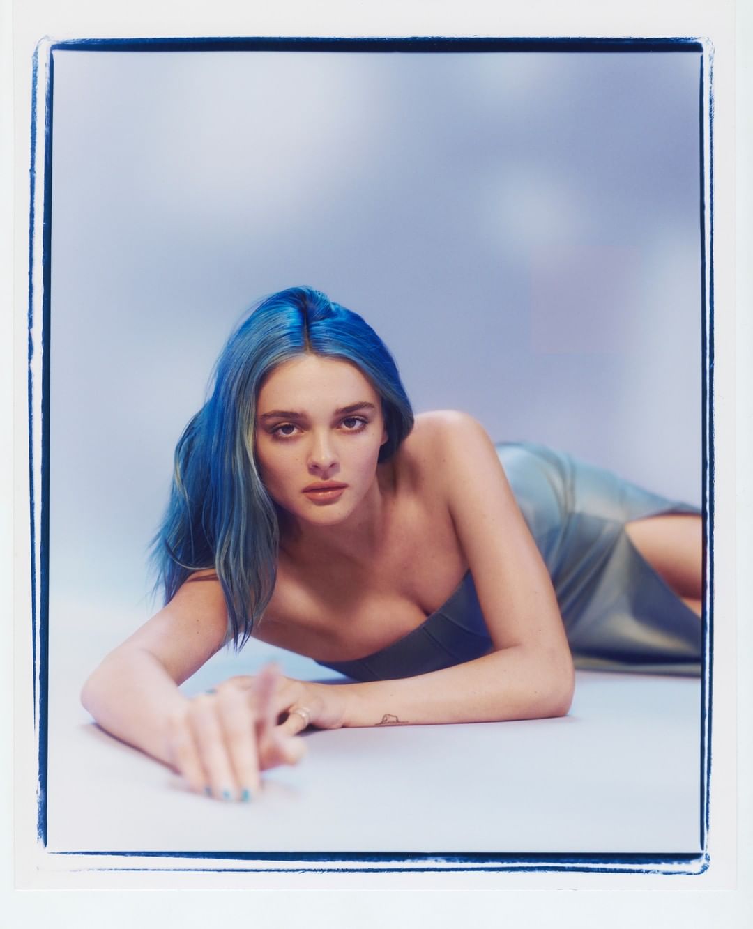 A young woman with blue hair and a strapless dress poses while lying down in a photograph with soft lighting and a cyanotype-style border.