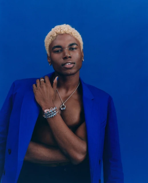 Portrait of a Black male model with blond hair wearing a blue jacket and jewelry against a blue backdrop in a studio setting.