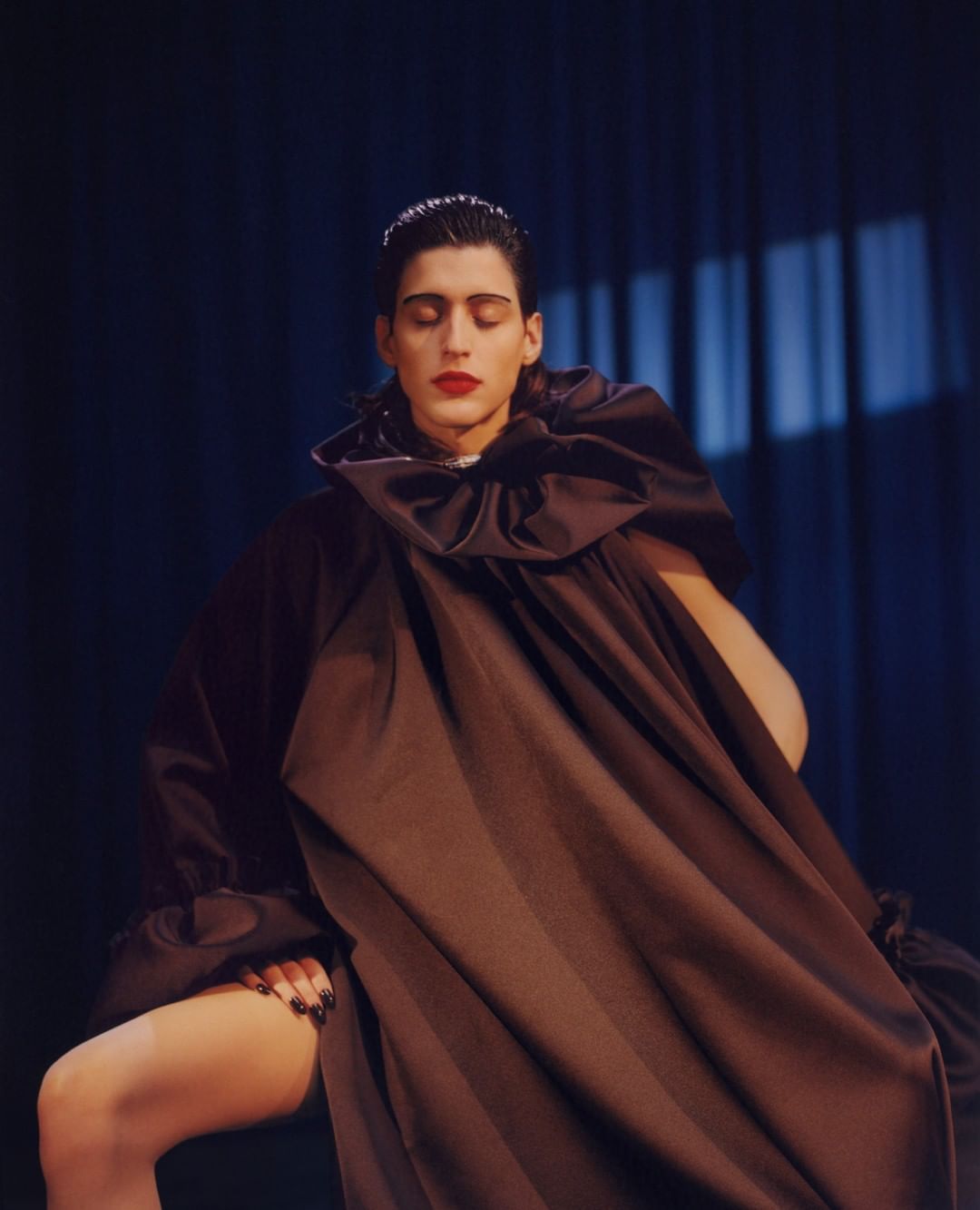 Fashion portrait of an adult male in an oversized brown satin gown, seated in a photo studio with blue curtains in the background.