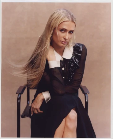 Blonde woman in a black dress with a white collar and cuffs poses in a chair against a neutral background. She rests her head on her hand.