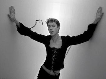 A model with short curly hair poses against a wall, wearing a black corset top and pants, with arms outstretched in a black-and-white studio shot.