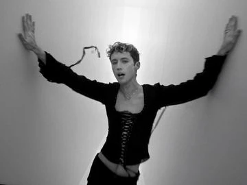 A model with short curly hair poses against a wall, wearing a black corset top and pants, with arms outstretched in a black-and-white studio shot.
