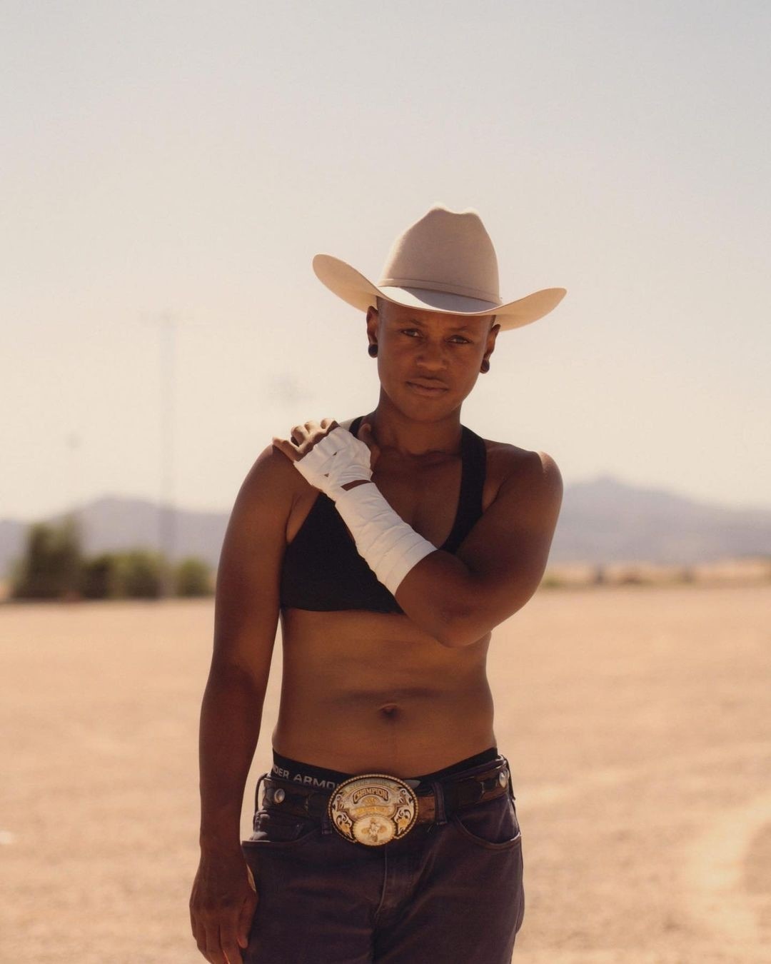 Portrait of a woman in a cowboy hat and boxing wraps stands in a desert landscape, wearing a black top and jeans with a large belt buckle.