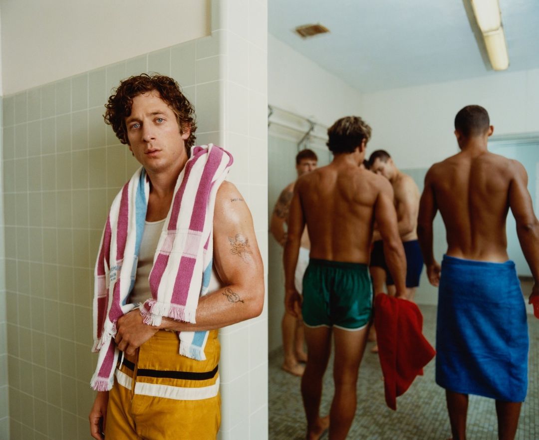 Portrait of a fair-skinned man wearing a striped towel and yellow shorts, plus a group of men in a locker room.