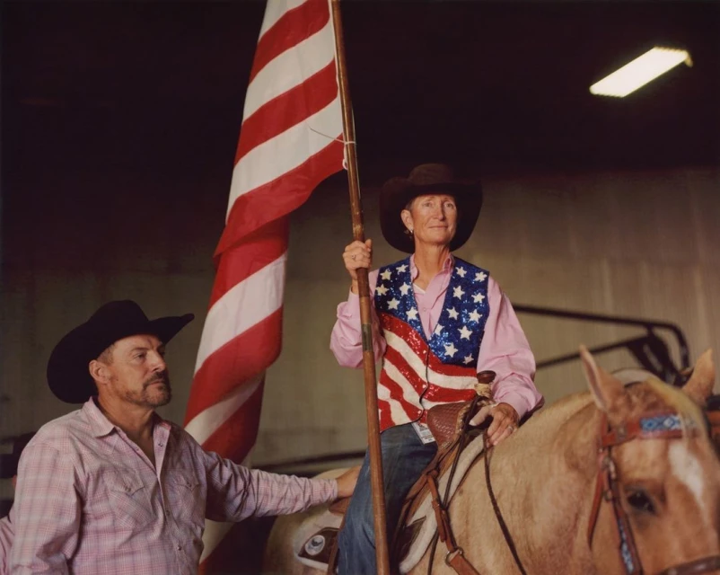 Two adults with a horse and US flag indoors. Woman in patriotic vest rides the horse as the man in a cowboy hat holds it.