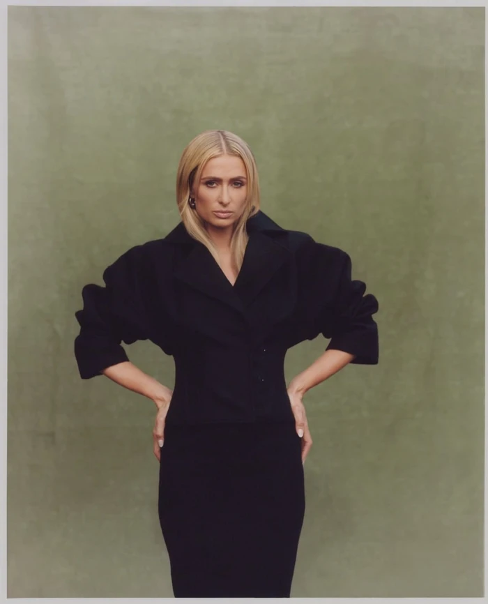 Paris Hilton with blonde hair wearing a black jacket and skirt against an olive green backdrop