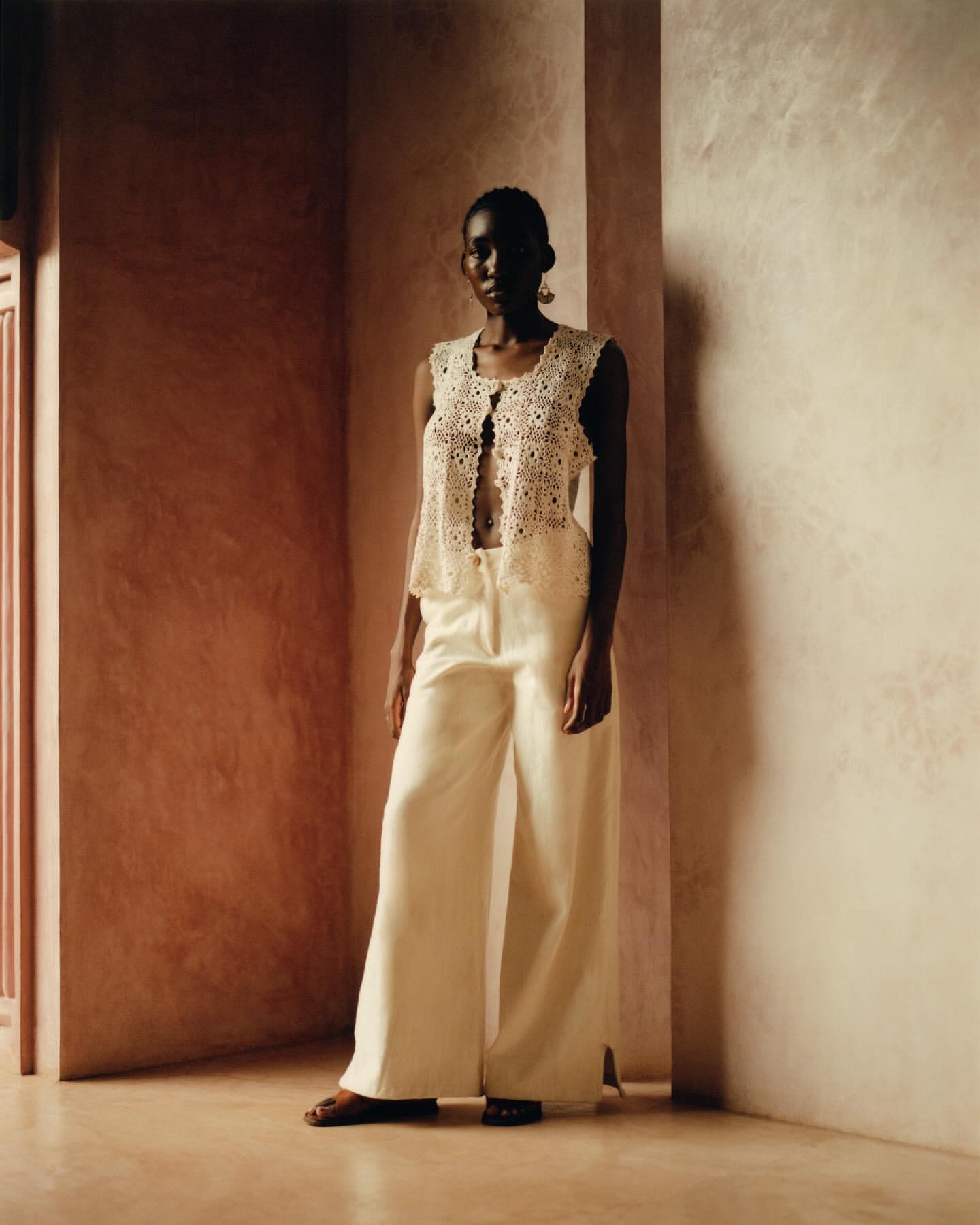 A slender African-American female model in a crochet vest and off-white wide-leg trousers poses in a brightly lit corner setting.