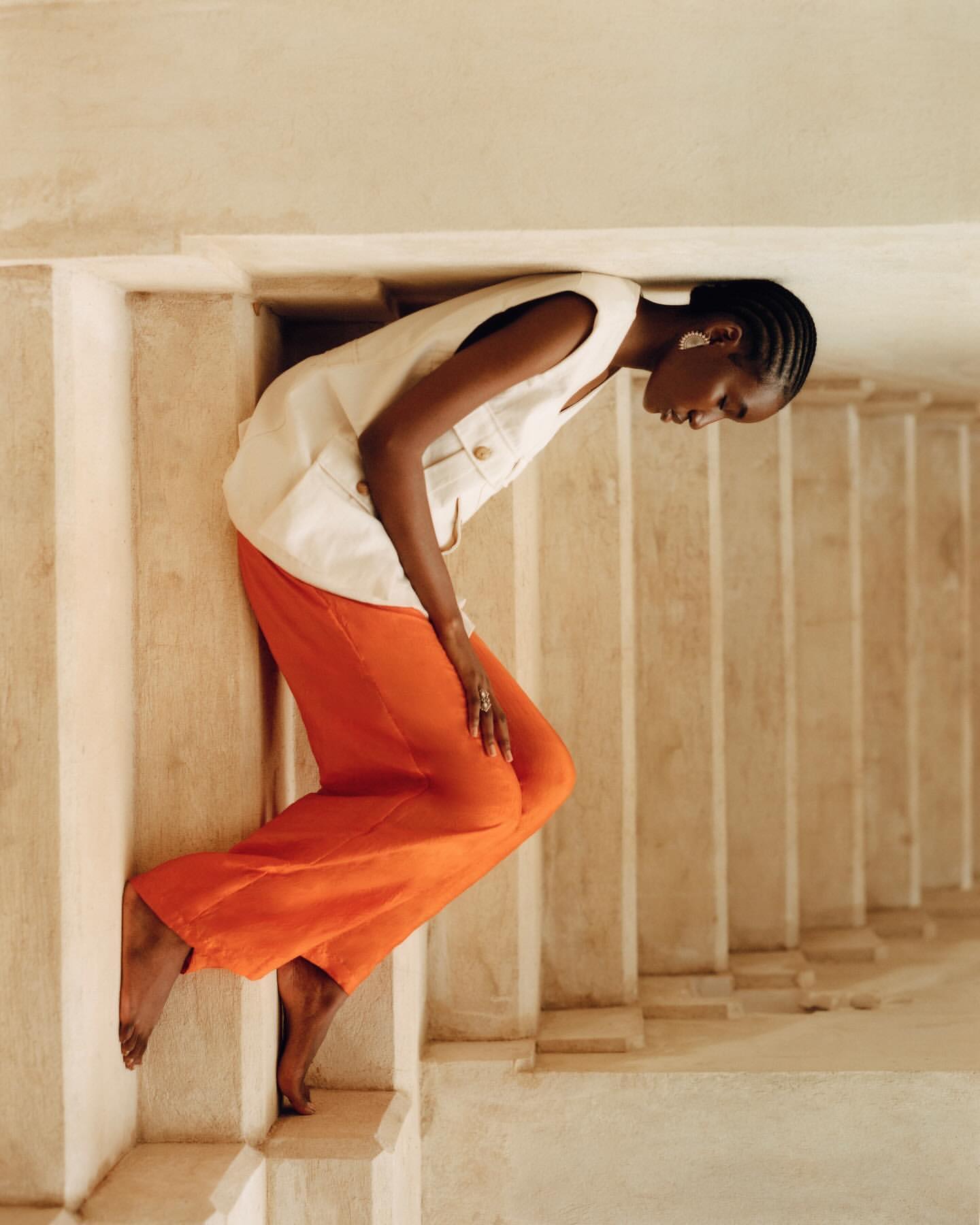 Fashionable woman poses barefoot on stairs wearing a white vest and orange linen pants in a minimalist indoor setting, lit by natural light.