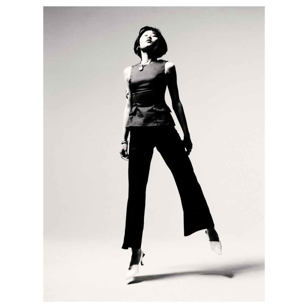 A woman in a dark top and pants strikes a pose in a studio setting, glancing up and away from the camera in black and white.