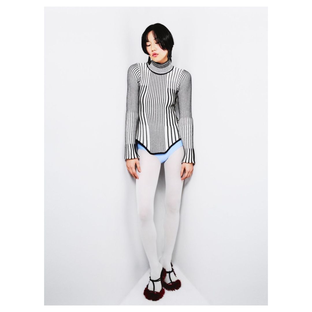 Young woman in studio posing with striped turtleneck sweater, white tights, and furry Mary Jane flats against a white corner wall.