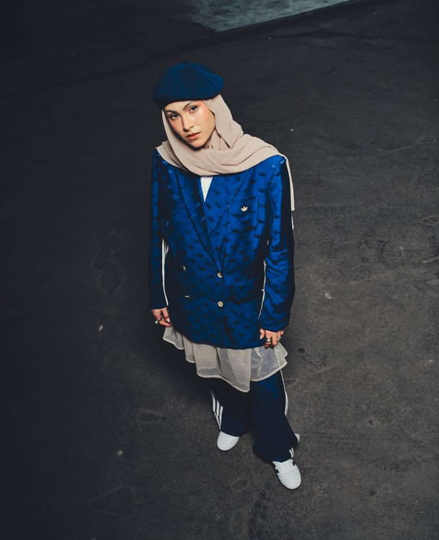 Woman in a blue blazer and headscarf poses in an urban setting. She also wears wide-leg pants and white sneakers, looking directly at the camera.