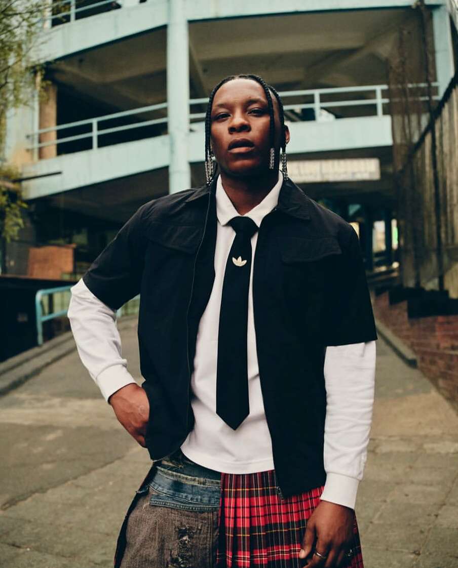 Stylish adult in black jacket, tie, and skirt stands outdoors against a concrete building, showcasing an editorial fashion portrait.