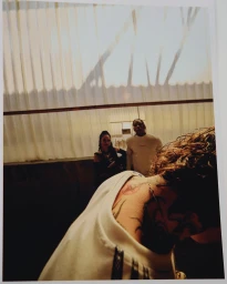 Polaroid photograph capturing a person in a white top facing away while two figures stand in the background near a light-filtered wall.