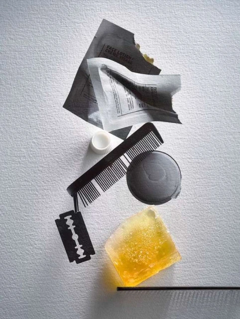 Still life composition featuring packets, a razor blade, soap, comb, bottle cap, and a cosmetic container on a textured white surface.