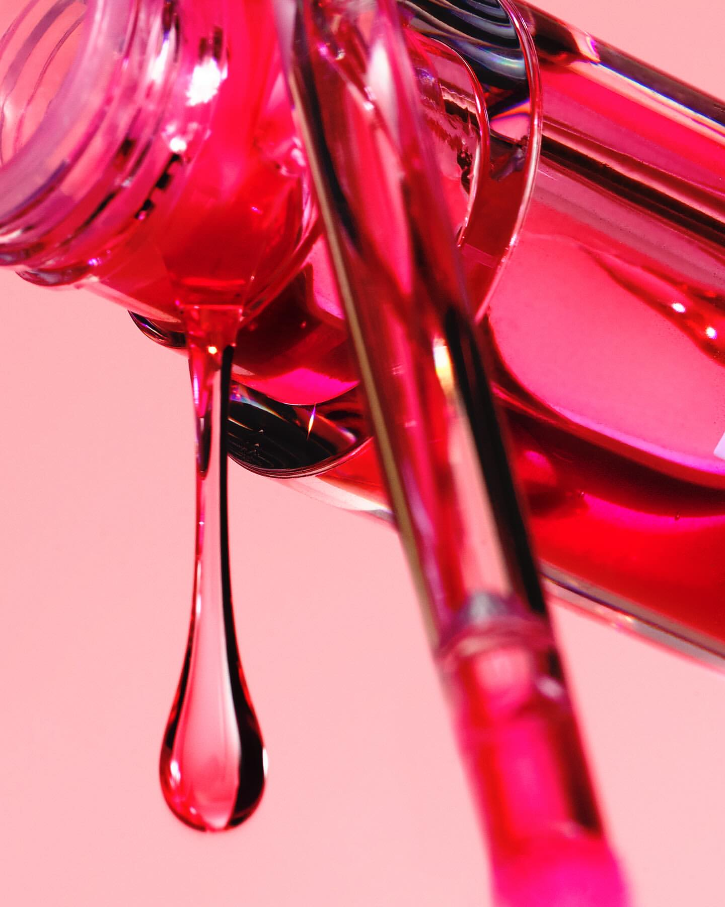 Macro shot of a tilted pink nail polish bottle with a brush and a large droplet of nail polish against a solid pink backdrop.