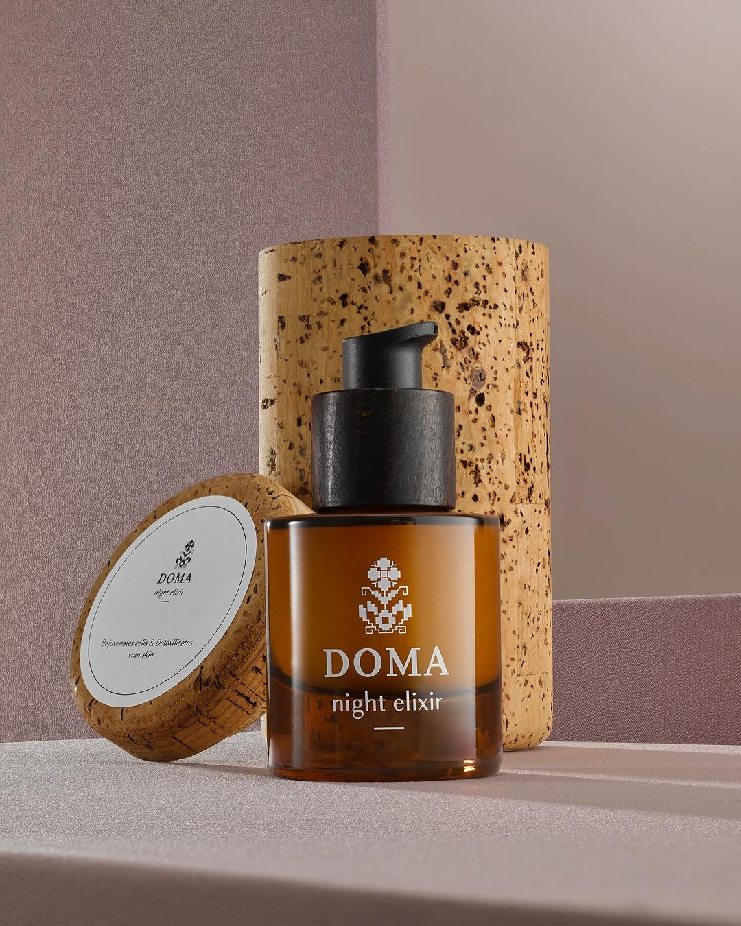 Amber-colored glass bottle of Doma night elixir with black pump, next to two cork coasters against a textured pink wall.