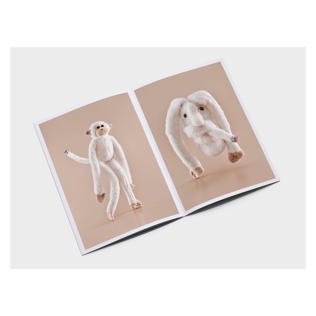 Open magazine spread features stuffed monkey and rabbit, both white, set against a neutral background, minimalist and whimsical.