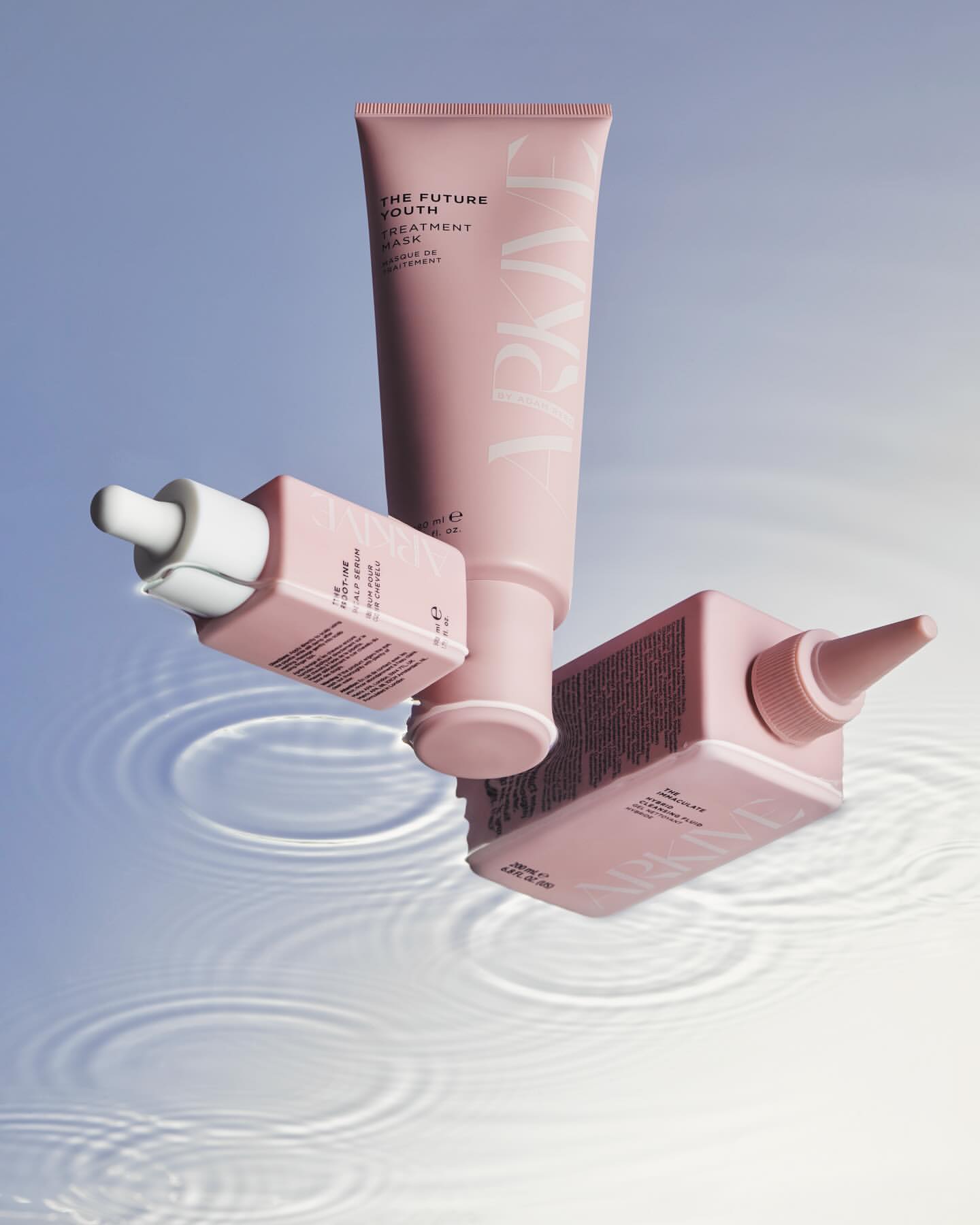 Three pink skincare products including a tube and dropper bottle sit on water with rippling effects. Cosmetic product shot for beauty brand.