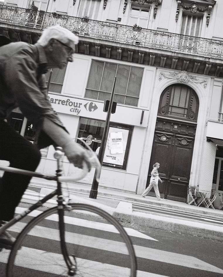 Monochrome street photography by Luke Kuisis of an elderly man riding a bicycle past a Parisian building facade.