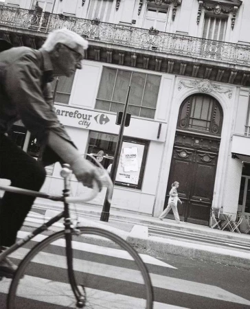Monochrome street photography by Luke Kuisis of an elderly man riding a bicycle past a Parisian building facade.