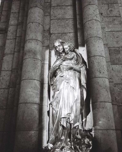 Monochrome statue of woman holding a child in an indoor environment, showcasing classical artistic style and stone architectural elements.