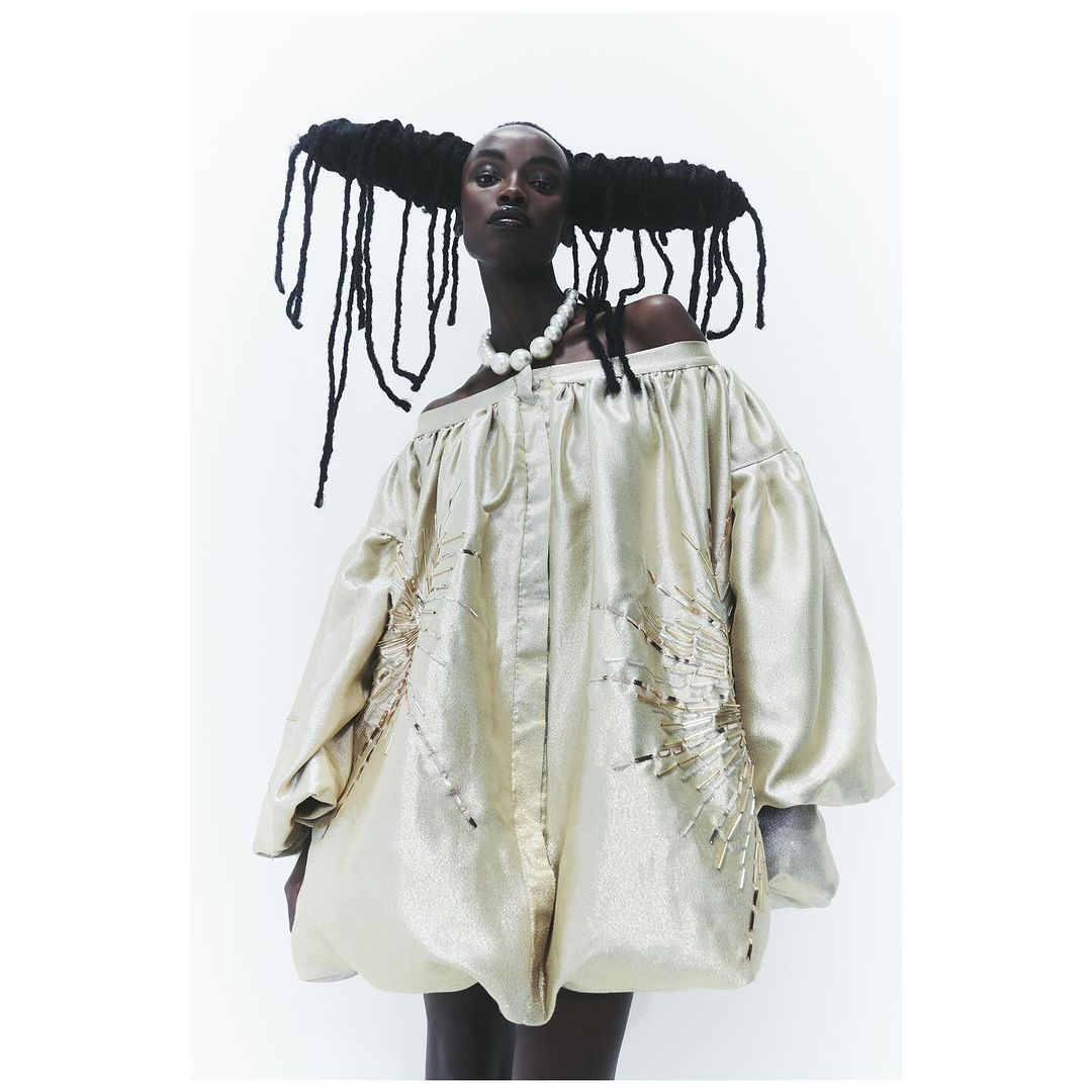 Portrait of black fashion model wearing pearl necklace and draped gold lame off-shoulder mini dress against plain white studio backdrop.