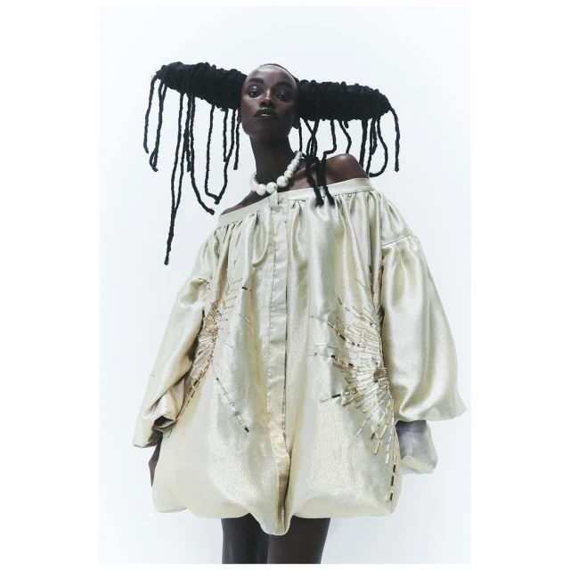Portrait of black fashion model wearing pearl necklace and draped gold lame off-shoulder mini dress against plain white studio backdrop.