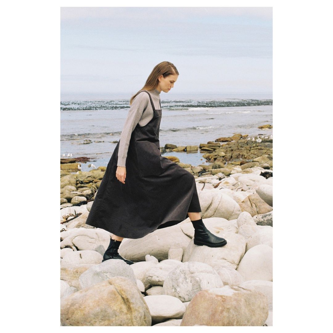 A woman in a dress and boots walks on rocks near a beach. The image captures a fashion moment in a natural, coastal setting.