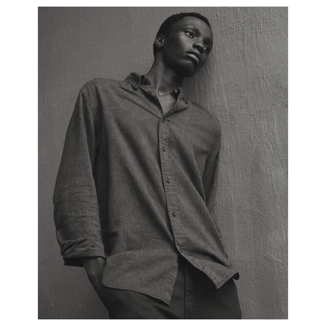 Monochrome portrait of a young African man in a button-down shirt leaning against a wall, eyes looking upward in a contemplative pose.