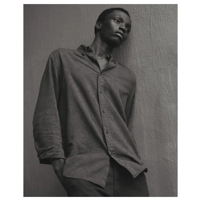 Monochrome portrait of a young African man in a button-down shirt leaning against a wall, eyes looking upward in a contemplative pose.