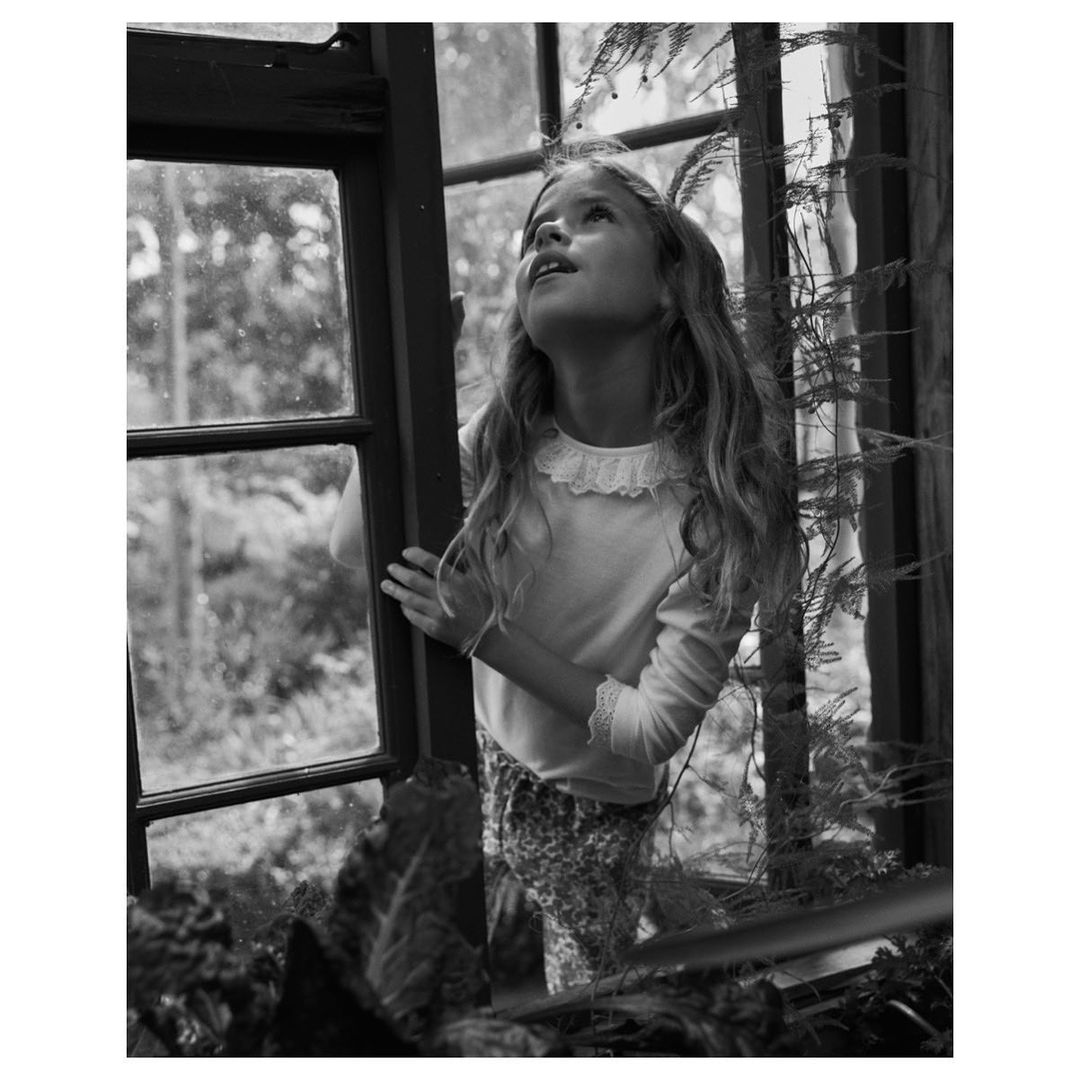 A young girl with long curly hair stands by a window, looking up, in a black and white photograph. The outdoor foliage is blurred.