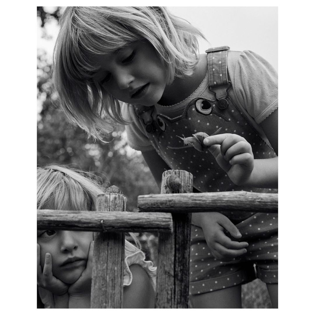 Two young girls are outside near a wooden fence; one girl is looking at a snail on her hand, and the other is resting her face on the fence.
