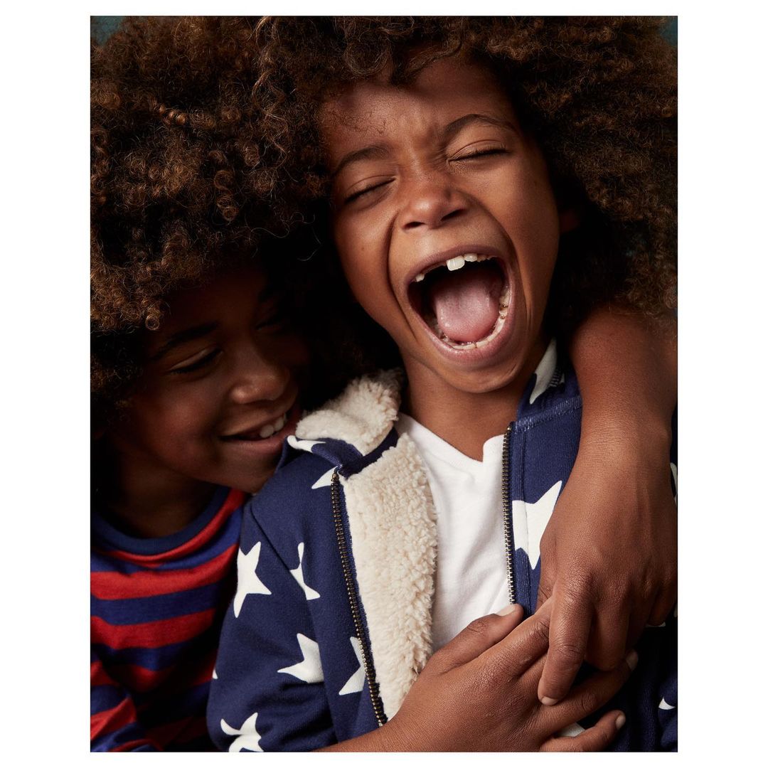 Two happy children hug; one child has their mouth open, eyes closed and the other child is smiling. They wear casual clothing.
