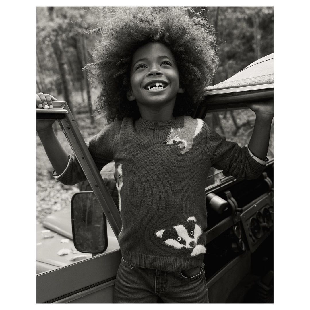 Black and white portrait of a smiling child in animal sweater, outdoors next to vehicle on a nature background, looking upward.