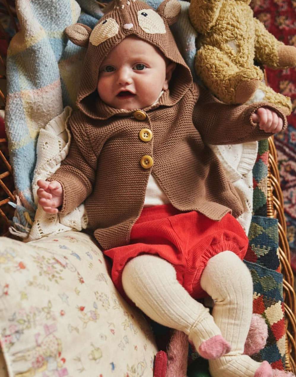 Baby wearing a knit fawn hooded sweater and red shorts is nestled in a wicker basket with pastel blankets and a stuffed teddy bear.