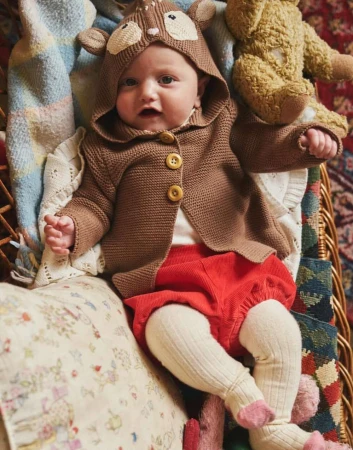 Baby wearing a knit fawn hooded sweater and red shorts is nestled in a wicker basket with pastel blankets and a stuffed teddy bear.