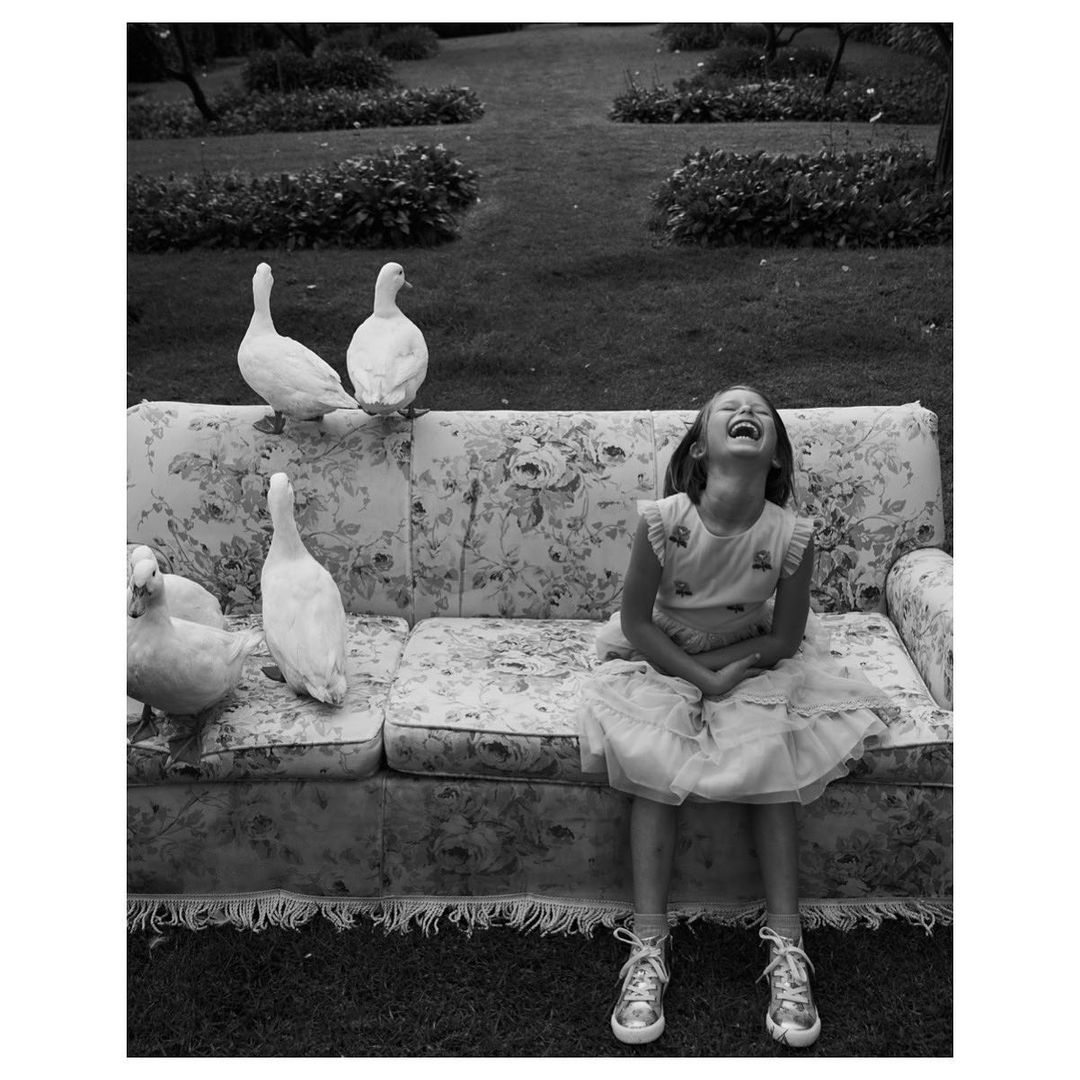 Black and white shot of a girl laughing on a floral couch surrounded by ducks in a yard, creating a whimsical and playful scene.