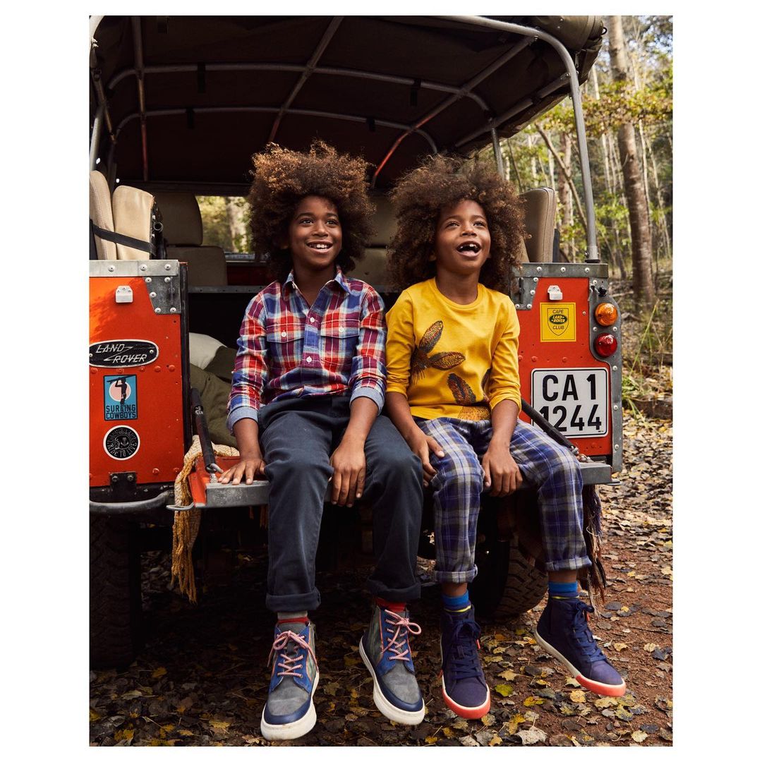 Two young boys with afro hairstyles sitting on the back of an orange Land Rover in a forest, smiling and looking up on a sunny day.