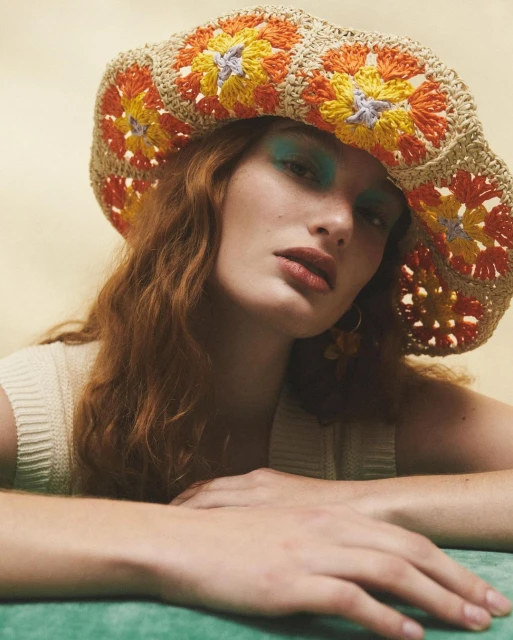 Portrait of a red-haired woman wearing a crochet flower hat, light makeup, and a knit top, posing in a neutral-toned indoor studio setting.