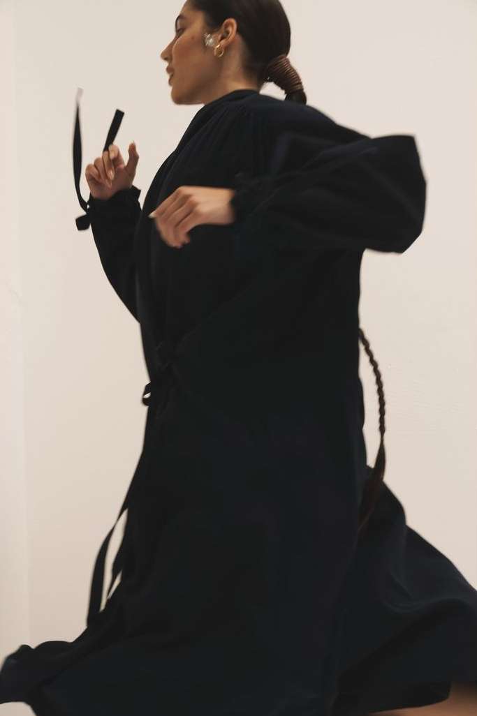 Woman in a dark flowing robe poses in motion against a light background, showcasing fashion and movement.