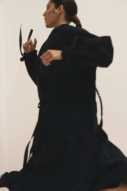 Woman in a dark flowing robe poses in motion against a light background, showcasing fashion and movement.