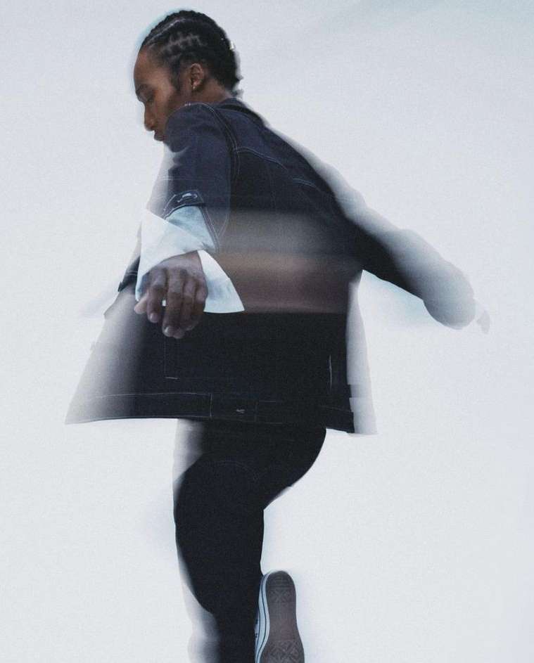 Side view of a blurred African American male in a black denim jacket and black jeans walking against a blurred white background.