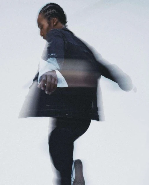 Side view of a blurred African American male in a black denim jacket and black jeans walking against a blurred white background.