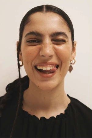 Close-up portrait of a smiling woman in a black top winking and sticking out her tongue against a plain neutral background.