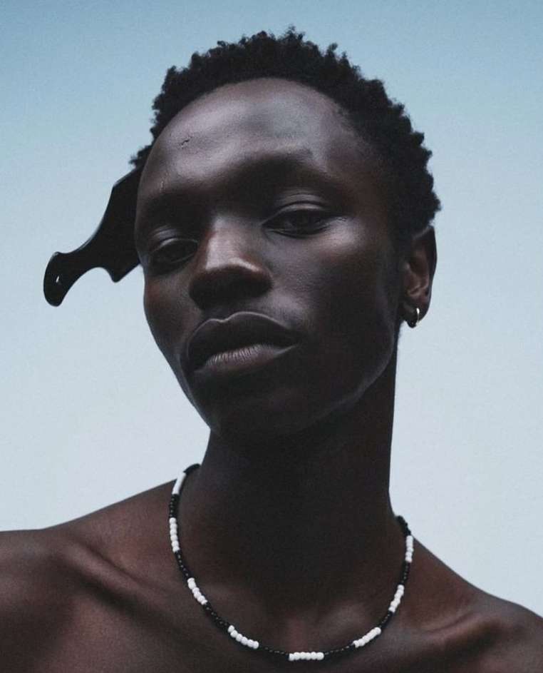 Close-up portrait of a dark-skinned adult male wearing a beaded necklace and a comb in his hair, against a light-blue background.