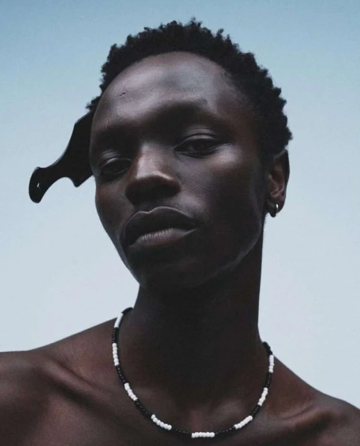 Close-up portrait of a dark-skinned adult male wearing a beaded necklace and a comb in his hair, against a light-blue background.