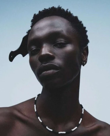 Close-up portrait of a dark-skinned adult male wearing a beaded necklace and a comb in his hair, against a light-blue background.