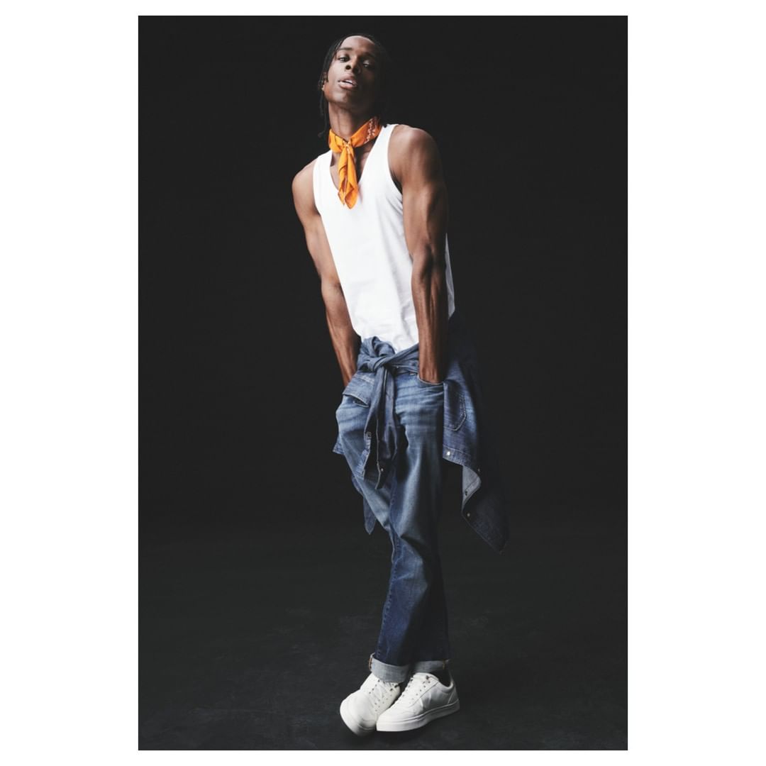 A slender adult male fashion model in a white tank top and jeans poses against a dark background in a studio setting with low-key lighting.