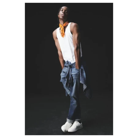A slender adult male fashion model in a white tank top and jeans poses against a dark background in a studio setting with low-key lighting.