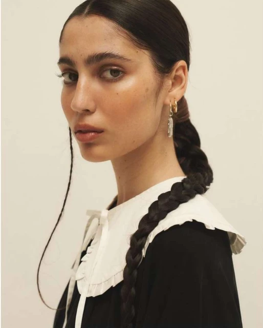 Portrait of a young woman with dark hair styled in a braid, wearing a black top with a white ruffled collar and dangling earrings.